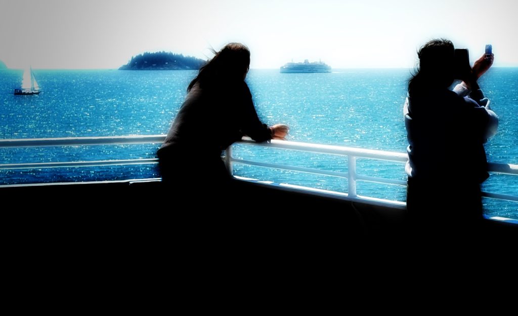 A person leans against the railing of a ferry while another person takes a selfie. A sailboat, an island, and another ferry can be seen in the background.